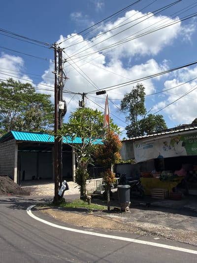 The image depicts a street scene in Tampaksiring, Indonesia, under a clear blue sky with scattered white clouds. A prominent utility pole with numerous wires crisscrosses the frame. To the left, a building with a bright blue corrugated metal roof and concrete block walls is visible. Adjacent to it is a pile of dark soil. In the center, a small tree with green and reddish foliage stands near a tall utility pole. Further to the right, another building is partially visible, featuring a banner with text and images, possibly a storefront. A motorcycle is parked in front of this building. A dark gray trash bin is situated on the sidewalk. The road in the foreground is paved, with a white line marking its edge. The environment appears to be a rural or semi-urban area, characterized by the presence of utility infrastructure, buildings, and vegetation.
