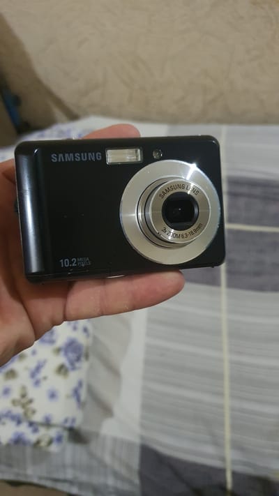 A black Samsung digital camera with a silver lens is held by a person's hand. The camera has "SAMSUNG" branding at the top and "10.2 MEGA PIXELS" near the bottom left. The lens has "SAMSUNG LENS" and "3x ZOOM 6.3-18.9mm" inscribed on it. The camera is positioned in front of a striped and floral patterned surface, possibly bedding or upholstery. The background is out of focus and appears to be a muted, textured wall. The image's lighting suggests an indoor setting. The image is not located in the city of Karankul, Uzbekistan.
