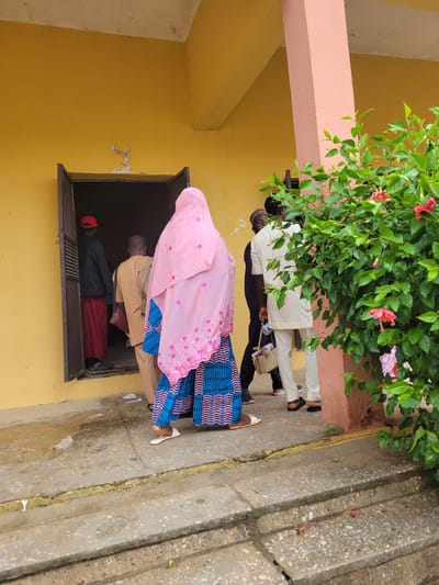 The image captures an outdoor scene in Keffi, Nigeria, depicting the entrance to a yellow-walled building with several individuals. The foreground features a weathered concrete pathway and two steps, with a faded yellow line marking the edge of the upper landing. Small pieces of white paper and plastic debris are scattered on the ground.
A dark, open doorway in the yellow wall is the central focus, with people either entering or queuing to enter. Visible through the doorway, a person wearing a red cap, dark jacket, and red trousers is moving further inside, with their back to the viewer. Directly in front of the doorway, a bald individual in light brown traditional attire with a dark shoulder bag is also facing away, appearing to enter.
In the immediate foreground, a person, likely female, is prominently featured from behind. They wear a light pink head covering adorned with embroidered patterns and a blue and pink patterned dress. White flat shoes are visible on their feet as they move towards the doorway.
To the right, partially obscured by a dense green bush with several red hibiscus flowers, stands another person in light cream/beige traditional tunic and trousers. This individual holds a light beige bag and possibly a white cup or container in their left hand, and wears a dark cap. Their back is to the viewer, facing towards the doorway. Behind them, a pinkish-peach colored pillar rises from the ground, framing the right side of the scene, with another individual partially visible in dark clothing. The building's ceiling, visible at the top, appears to be a simple, light-colored structure. The scene is illuminated by natural daylight.