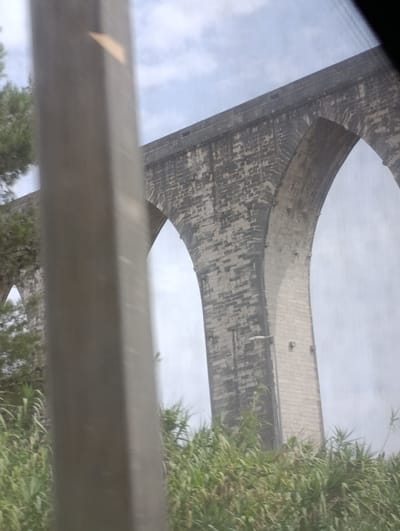 The image shows a portion of the Aqueduto das Águas Livres in Lisbon, Portugal. A tall, grey, rectangular structure is prominently in the foreground, slightly out of focus and obscuring part of the background. Beyond this structure, a section of the aqueduct's stone arches and masonry is visible against a cloudy sky. The aqueduct is old, with visible weathering on the stone. Lush green foliage, likely trees and bushes, is seen at the base of the aqueduct and in the foreground. The lighting suggests it is daytime, possibly overcast. The overall scene captures a fragment of historic architecture surrounded by vegetation.