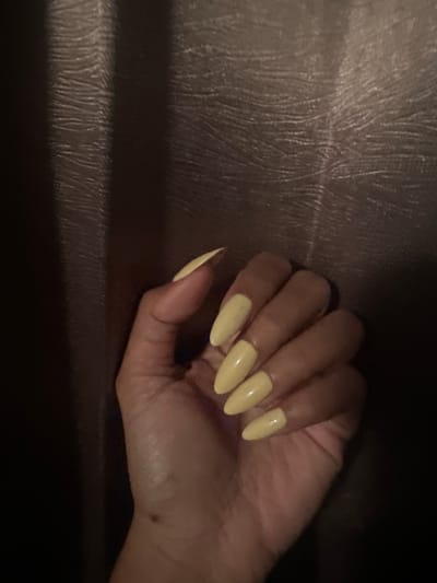A left hand with long, almond-shaped fingernails painted a pale yellow is held up against a textured, dark brown background. The hand is the primary subject of the image, with the nails catching the light and appearing glossy. The background appears to be fabric, possibly a curtain or upholstery, with a ribbed texture. The lighting is dim, casting shadows and creating a moody atmosphere. There is no discernible location within Sukawati, Indonesia, provided by the image itself.
