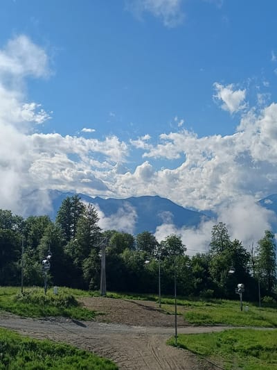 The image depicts a mountainous landscape with a clear blue sky filled with white, fluffy clouds. In the foreground, a dirt path winds through green grassy slopes, with several ski lift towers and chairs visible among the dense green trees that line the mountainside. In the background, majestic mountains rise, partially shrouded in mist and clouds, suggesting a high altitude. The scene appears to be an outdoor recreational area, likely a ski resort during the off-season, given the presence of ski lifts and the lush greenery. The location is identified as Estosadok, Russia.
