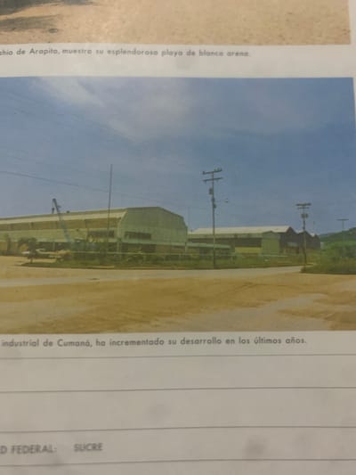 This is a photograph of a landscape that appears to be a beach with industrial buildings in the background. The image is oriented vertically. In the foreground, on the left, there is text written vertically, describing the "playa de blanca arena" (white sand beach) of Arapito. The middle ground shows a large, light-colored industrial building with a white roof, possibly a warehouse or factory. There are also utility poles with wires running across the sky. In the background, there are hills covered in green vegetation. The sky is clear and blue, suggesting it is daytime with good weather. Further text is present on the right side of the image, also oriented vertically, which states "industrial de Cumaná, ha incrementado su desarrollo en los últimos años" (industrial of Cumaná, has increased its development in recent years) and also mentions "D FEDERAL: SUCRE". The overall scene depicts a coastal area that is undergoing industrial development. There are no people visible in the photograph.