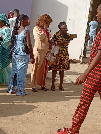 The image displays an outdoor scene in Sabon Gari West, Nigeria, under bright daylight, with strong shadows indicating sunlight. The ground is unpaved and appears to be light brown dirt or sand. In the background, a light-colored building wall with vertical metallic paneling is visible, along with a doorway and steps further back, where additional individuals are present.

Five primary subjects are in the foreground and midground:
1.  On the far left, partially obscured, a female wears a green patterned headwrap and dress, holding a dark object.
2.  A female with dark skin and long, dark braided hair tied in a high ponytail walks towards the left, wearing a light blue lace two-piece outfit (top and wide-leg pants) and pink-rimmed sunglasses. She carries a black shoulder bag.
3.  In the center, a female with dark skin and light brown curly hair wears a long, light tan dress with a red inner lining visible. She has a watch on her left wrist and a small, woven metallic clutch bag hanging from her left shoulder. She appears to be conversing with the person to her right.
4.  To her right, a female with dark skin and dark hair styled back with a headband wears a yellow dress with a black geometric pattern. She holds a black smartphone in her left hand and gestures outwards with her right hand. She carries a white tote bag with black text and another small, woven metallic bag.
5.  In the immediate foreground on the right, the lower body of an individual with dark skin is visible, walking towards the left. This person wears brown pants with a red zigzag pattern and red sandals.

Several other individuals are partially visible in the background, including a male in a white shirt behind the braided-hair female, and others near the building entrance dressed in light blue and green attire.