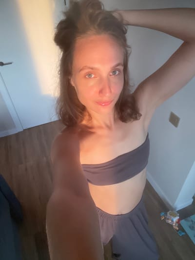 A woman is captured in a selfie, lying down and holding her arm above her head. She is wearing a dark grey sports bra and matching grey sweatpants. Her eyes are open and looking towards the camera, and her lips are slightly parted, with a hint of a smile. The lighting suggests it is either sunrise or sunset, casting a warm glow on her skin. The background includes a white wall, a wooden floor, and a door. A small, colorful toy is visible in the upper right corner of the image, suggesting the presence of a child. The overall mood is relaxed and intimate, with a touch of warmth from the lighting. The location is identified as Podshivalovo, Russia. There is no visible text in the image.