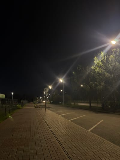 The image depicts an outdoor scene at night, likely in Naberezhnye Chelny, Russia. A paved walkway, made of brick-like material laid in a repeating pattern, occupies the foreground and extends into the distance. Adjacent to the walkway is a parking lot with visible white parking space lines. On the right side of the image, a dense tree with bright green foliage is illuminated by a streetlamp, creating a strong glare that casts light rays. Several other streetlights are visible throughout the scene, casting pools of light and illuminating the pathway and surrounding areas. The sky is dark, indicating nighttime. In the background, there are hints of other structures and possibly more trees. The scene appears to be an urban or suburban area, possibly a park, a pedestrian zone next to a road, or an area adjacent to a commercial or residential complex. There are no people or vehicles explicitly visible in the frame.