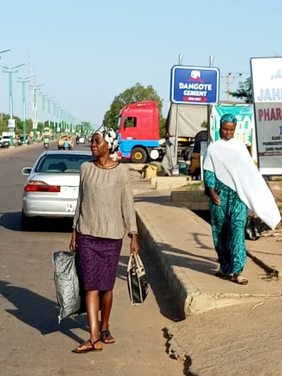 Two women are walking on the side of a street in Tumfure, Nigeria. The woman on the left is wearing a gray long-sleeved shirt and a purple patterned skirt. She is carrying a large, light-colored bag in her left hand and a black and white tote bag in her right hand. She is wearing brown sandals. The woman on the right is wearing a teal patterned dress and a white headscarf draped over her shoulders. She is wearing light-colored sandals.
In the background, a silver car is parked on the left side of the road. A red and blue truck is visible further down the street. There is a blue sign with white lettering that says "DANGOTE CEMENT". A white sign with red lettering that says "JAHI PHARMA" is also visible. The street is lined with streetlights on poles. The ground is paved, and there is a concrete curb on the right side of the street. The sky is clear and blue.