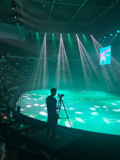 A cameraman stands on a platform in an arena, filming an ice rink illuminated with green lights and a projected pattern. The cameraman is silhouetted against the bright ice. In the background, tiered seating for spectators is visible, with some audience members in the lower sections. Several spotlights shine down from the ceiling, creating beams of light that cut through the smoky atmosphere. A large screen at the far end of the arena displays colorful graphics and text in Russian. The scene appears to be set up for a performance or event. The location is the Platinum Arena in Khabarovsk, Russia, a multi-purpose indoor arena.