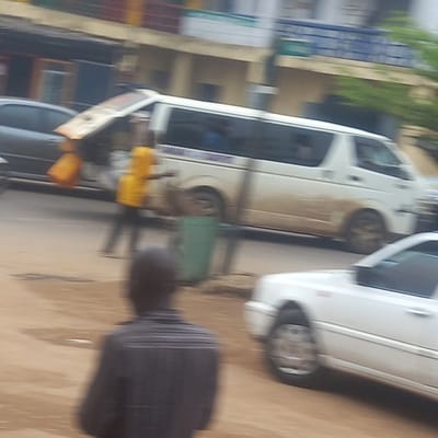 The image, taken in Sardauna, Nigeria, shows a street scene with a white van splattered with mud parked alongside a white car. Two people are vaguely visible near the van. A person in a dark striped shirt stands in the foreground. The background includes a building and hints of foliage. The street surface appears to be brown.