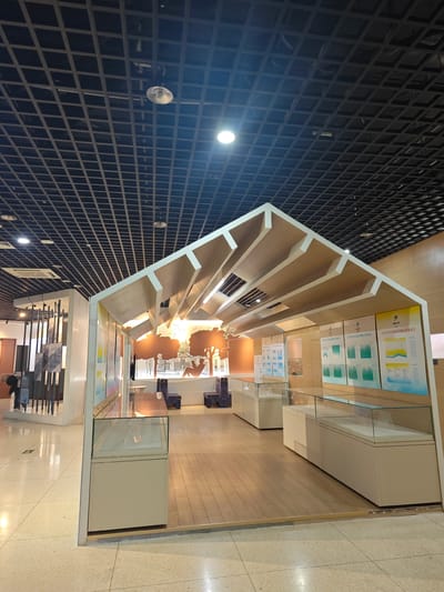 The image captures an indoor scene, likely within a museum or exhibition space located in Daxing District, China. A prominent feature is a booth structure resembling a house frame, constructed from light-colored wood with white accents. The roof section has a unique design, featuring alternating solid beams and open, square spaces.

Inside the booth, there are display cases, also in light colors with glass tops. The cases appear to be showcasing exhibits, though the specific contents are not fully visible. Walls behind the cases feature mounted posters, including some with Chinese text.

The surrounding environment includes a polished floor with a mottled pattern, and a suspended ceiling grid with recessed lighting. A blurred figure can be seen near a metal-barred structure in the background, suggesting the presence of other exhibits. The lighting is bright and even.
