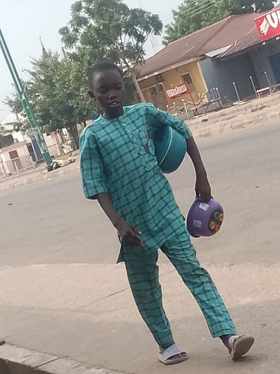 The image depicts a young boy walking on a sidewalk in Pantami, Nigeria. He is wearing a teal patterned traditional outfit and carrying a light blue bowl in his left arm and a purple container with colorful inserts in his right hand. The background shows a street scene with buildings, trees, and a traffic light pole. The boy is the central subject of the image, and his movement suggests he is walking somewhere. The environment appears to be an urban or semi-urban area with typical street infrastructure.