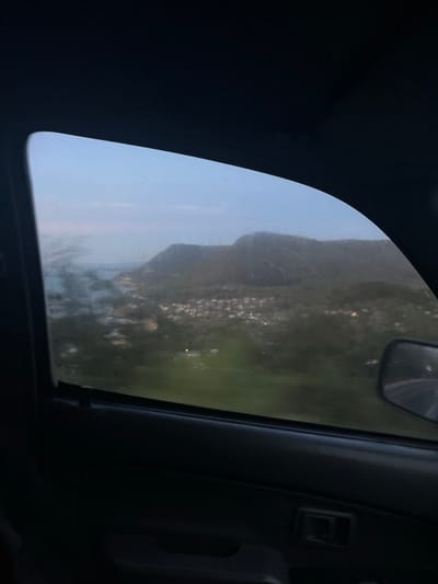 The image, taken from inside a vehicle, shows a view of Stanwell Tops, Australia through a side window. The landscape includes a mountain or hill and a body of water, possibly the ocean. There are faint lights visible, indicating a town or settlement in the distance. The interior of the vehicle is dark, with portions of the dashboard and the side mirror visible. The window glass appears slightly smudged or scratched.