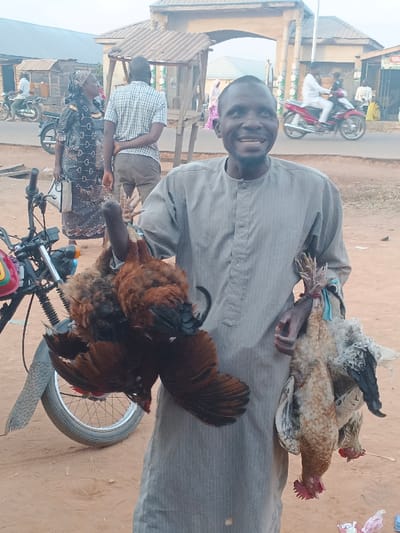 A man in a grey robe smiles holding chickens. He holds two brown chickens in his left hand and two white chickens in his right. Behind him are other people, motorcycles, and buildings. The scene appears to be outdoors in a rural area or small town. The lighting suggests it could be early morning or late afternoon.
