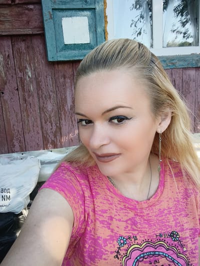A woman with blonde hair, wearing a pink and orange t-shirt and a silver earring, poses for a selfie. She is standing in front of a weathered wooden wall with peeling paint. Behind her, there is a window with a rustic wooden frame and a white pane. To the left, white plastic bags are visible. The scene appears to be outdoors, possibly in a rural setting or an older part of a city like Chernihiv, Ukraine. The lighting suggests it is daytime.