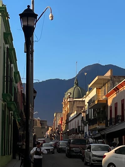 The image depicts a street scene in Oaxaca City, Mexico. In the foreground, a black, ornate street lamp stands out against a bright blue sky. A green-roofed building is partially visible beneath the lamp. In the midground, a domed building is situated in front of a large mountain. Further down the street, people are visible on the sidewalk. Parked cars line the right side of the street. The buildings lining the street appear to have colonial architecture with various colors. The light suggests it is either morning or late afternoon.