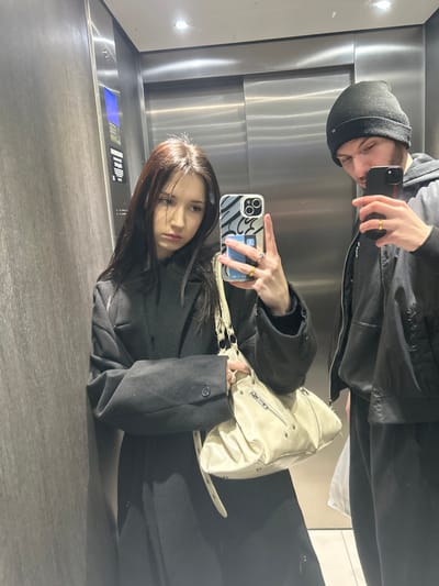 Two people in an elevator take a mirrored selfie. One, wearing a black beanie, holds a phone. The other, with long brown hair and a black coat, also holds a phone and a light-colored shoulder bag. The elevator has stainless steel walls.