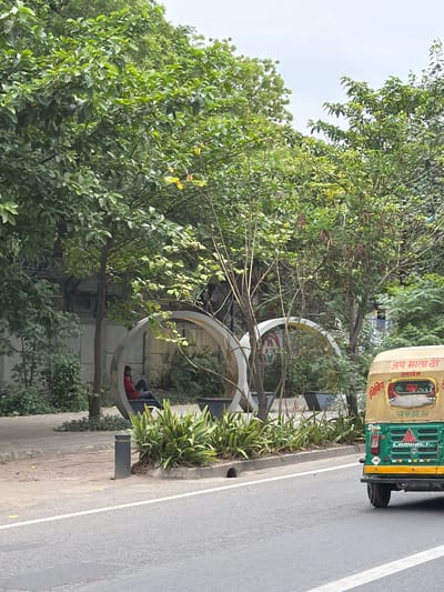 The image shows a street scene in New Delhi, India, with lush green trees lining a sidewalk. In the foreground, a yellow and green auto-rickshaw is partially visible on the right side of the frame, with text in Hindi on its rear. The auto-rickshaw has "Jay Mata Di" written on its top and "Compact" on its back. On the sidewalk, two modern, circular seating structures are visible. One structure has a person in a red shirt sitting inside. A bollard stands on the sidewalk next to a bed of green plants. The street in front of the sidewalk is asphalt with white lane markings. The sky is overcast.