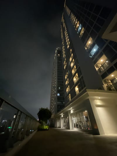 The image depicts a nighttime view looking up at a modern skyscraper in Central Jakarta, Indonesia. The building is illuminated, with many windows lit, suggesting it is occupied. The perspective is from a lower vantage point, looking upwards and slightly across the building's facade.
The scene is set at night, as indicated by the darkness and the artificial lighting of the building and the city below, which is reflected on the wet-looking ground in the foreground. The weather appears to be misty or foggy, obscuring the view of the lower parts of the city and creating a soft backdrop for the illuminated building.
In the foreground, there is a balcony or elevated walkway with a glass railing. Beyond the railing, a small, well-maintained tree is visible, as well as what appears to be another section of the building or an adjacent structure with more lights. The overall mood is one of urban sophistication and the quiet energy of a city at night. No people are clearly visible, and no text is present in the image.