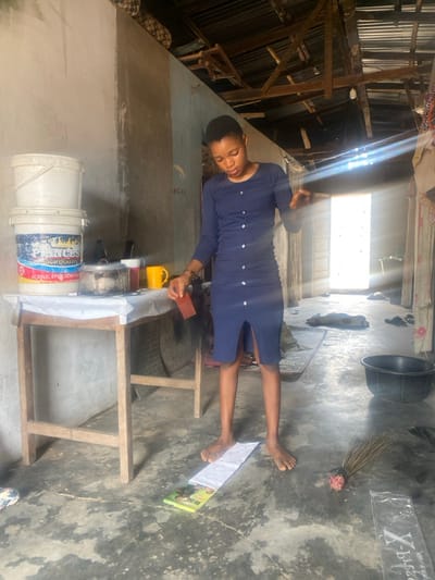 The scene is set indoors in Idiroko, Nigeria. A barefoot individual, wearing a navy blue, long-sleeved dress with a row of light-colored buttons down the front and a lower front slit, stands centrally. They are looking down, focused on an activity below eye level, holding a small, reddish-brown rectangular object in their right hand. Their bare feet rest upon a narrow white cloth strip laid on the mottled gray concrete floor, with one foot also placed over a small, colorful booklet or magazine.
To the left, a simple wooden table is draped with a white cover. On the table, two white plastic buckets are stacked, the lower one labeled "PRINCES HIGH QUALITY ACRYLIC EMULSION PAINT." Adjacent to the buckets are a lidded metal pot, a small dark container, and a yellow ceramic mug.
The room features unpainted, light-colored concrete walls and an exposed ceiling structure revealing wooden rafters supporting corrugated metal roofing sheets. Natural light streams in from an open doorway in the background, creating distinct sunbeams. On the concrete floor, a traditional broom lies near the bottom right corner, alongside a partial clear plastic sheet with "X-PIECES" text. Further back, a black plastic basin and various dark objects are visible on a mat near the doorway.