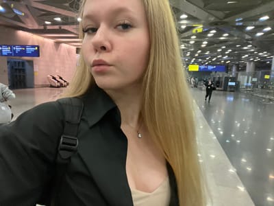 The image captures a young blonde woman taking a selfie in what appears to be a modern airport terminal. She is wearing a black button-up shirt over a beige top and a delicate necklace. Her hair is long and blonde, partially obscuring the background. The airport environment features a distinctive ceiling with triangular light fixtures and a polished floor reflecting the lights. In the background, signs in Cyrillic script indicate directions to "STORAGE" and "ARRIVAL". Other individuals are visible in the distance, one in a dark uniform walking, and others at a check-in or security area. A black and white patterned suitcase is also visible near the left side of the frame. The overall setting is clean and contemporary, consistent with an international airport like Vnukovo International Airport in Moscow, Russia.