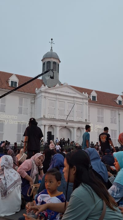 The image depicts a scene in front of the Gouverneurskantoor building, located in the Fatahillah Square of the Kota Tua area in West Jakarta, Indonesia. A large crowd of people is gathered outdoors, sitting on the ground, suggesting an event or performance is taking place. In the background, the white colonial-era building with a distinctive dome and clock tower stands prominently. A stage with musical equipment and performers is visible in front of the building. A large camera crane is positioned on the left side of the frame, indicating that the event is being filmed. The foreground features a group of people, including several women and a young boy, some of whom are wearing headscarves. The overall atmosphere appears to be relaxed and communal, with people enjoying the outdoor gathering.