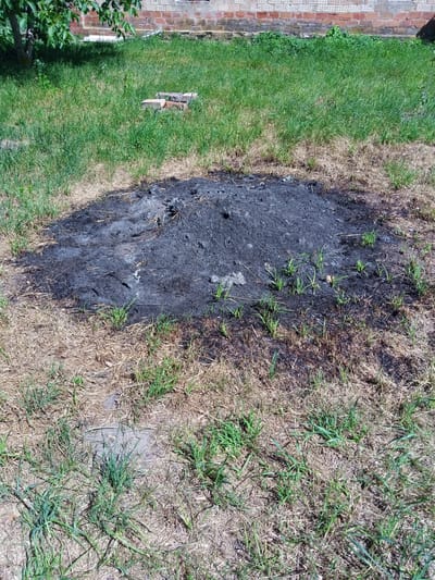 The image depicts an outdoor scene in Luka, Ukraine, likely during daylight on a sunny day. The primary focus is a large, dark pile of ash and charred debris in the center of the frame, suggesting a recent fire. The ash mound is surrounded by dry, yellowed grass, with patches of greener grass sprouting around its edges and further out into the yard. In the background, a brick wall is visible, possibly part of a building. To the left, a portion of a tree with green leaves casts a shadow over the grass. Several red bricks are scattered on the grass to the left of the ash pile, adding to the sense of disarray. The overall impression is one of a backyard or unkempt area that has experienced a fire, with nature slowly reclaiming the space. There is no visible text in the image.