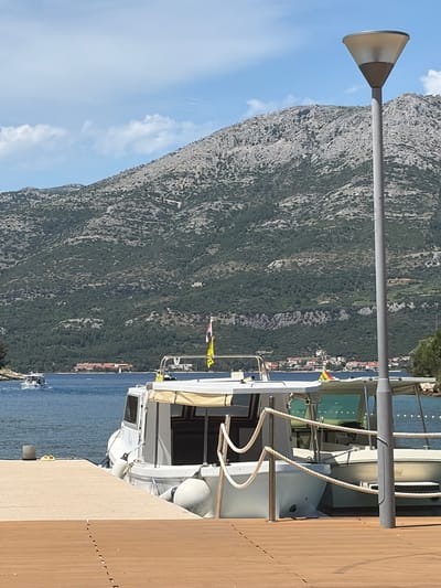 The image captures a sunlit coastal scene in Grad Korčula, Croatia. In the foreground, a light brown wooden pier extends from the bottom right, meeting a light concrete pier on the left. A pair of yellow flip-flops rests on the concrete pier near a metal bollard. Two white motorboats are docked side-by-side along the concrete pier, secured by ropes and multiple white fenders. The closer boat features a cream-colored canopy and displays a small Croatian flag and a yellow flag. A rope railing spans between the pier and this boat. Dominating the right side of the foreground is a tall, grey cylindrical street lamp with a conical, light-colored fixture.
The midground features calm, blue-green water. In the distance on the left, a third white motorboat moves across the water, generating a faint wake. Across the bay, a mountainous landscape forms the background. The mountains are rugged, characterized by a mix of grey rock and dark green, sparse vegetation. At the base of the distant mountains, a cluster of buildings with red-tiled roofs is visible along the shoreline. The sky above is clear light blue with scattered white cumulus clouds.