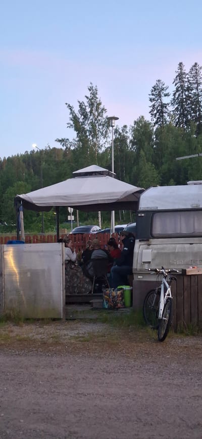 In Salpakangas, Finland, a group of individuals is gathered under a white gazebo. The gazebo is situated outdoors, adjacent to a light-colored caravan. Several people are seated around a table, appearing to be engaged in conversation or eating. A white bicycle is parked to the right of the caravan. The environment appears to be a wooded area with a clear sky overhead, possibly during twilight. The ground is covered in gravel and some vegetation.