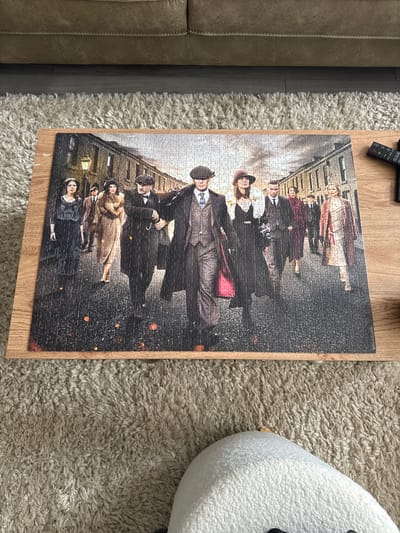 A framed puzzle depicting a scene from the television series Peaky Blinders is laid out on a wooden table. The puzzle features a group of characters, dressed in period clothing, walking down a cobblestone street lined with brick buildings. Several figures, including men in hats and suits, and women in dresses and fur coats, are prominently displayed. The street appears to be lit by a gas lamp. The puzzle is positioned on a wooden table which is placed on a shaggy, beige carpet. In the background, a portion of a beige sofa is visible. The scene is brightly lit, likely by natural light from a window or overhead lighting. The puzzle is oriented horizontally. There are no discernible elements that place the scene specifically in Craigavon, United Kingdom; the setting is consistent with a period drama.