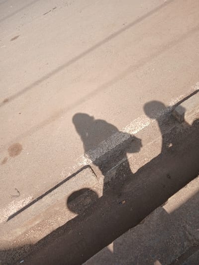 The image displays a ground-level perspective of a light-colored asphalt road or paved surface, exhibiting minor stains and aggregate details. A white, dashed line is painted diagonally across the road. To the right, a concrete curb defines the edge of the road, bordering a dark, recessed gutter or drainage channel containing some debris. Prominent shadows of at least two human figures are cast onto the road, over the white line, across the curb, and into the gutter. One shadow, appearing to be a person, is positioned near the white line, with an indistinct shape in its right hand. The long, sharp definition of the shadows indicates strong sunlight and a low sun angle. The scene depicts a public thoroughfare, and the specific location of "Hospital ward, Kenya" is not identifiable from the visual content.