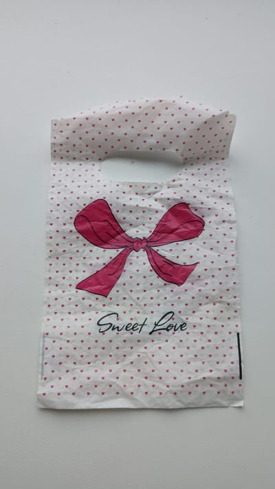 This media file shows a white plastic bag with a repeating pattern of small pink hearts. A large, pink bow with black outlines is printed in the center of the bag. Below the bow, the words "Sweet Love" are written in cursive black text. The bag appears to be a gift bag. The bag is lying flat against a white surface, likely a table, with a light-colored wall in the background. The lighting is even and appears to be from an overhead source. The overall impression is of a simple, decorative item. There is no activity taking place other than the bag being displayed. The location context provided is Kapyĺ, Belarus.