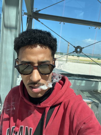 A young male subject with dark, curly hair and a distinct mustache is depicted in a close-up image, exhaling a plume of white vapor or smoke from his mouth. He wears tortoiseshell-patterned sunglasses, a red hooded sweatshirt over a black turtleneck, and a black strap of a bag diagonally across his chest. The subject is situated within a modern, glass-enclosed structure, likely an elevated walkway or viewing area, as evidenced by visible grey metal support beams, cables, and connectors for the large glass panels.
Through the transparent panels, a bright, clear blue sky with minimal white clouds is visible. The background reveals a coastal environment, featuring a light-colored terrain, possibly sand or dry vegetation, a concrete or paved walkway, and a metallic railing. Beyond these elements, a significant body of water, consistent with the sea, extends to a distant coastline or landmass. The architectural style and coastal view are consistent with a location like the port or a modern waterfront development in Tanger, Morocco.