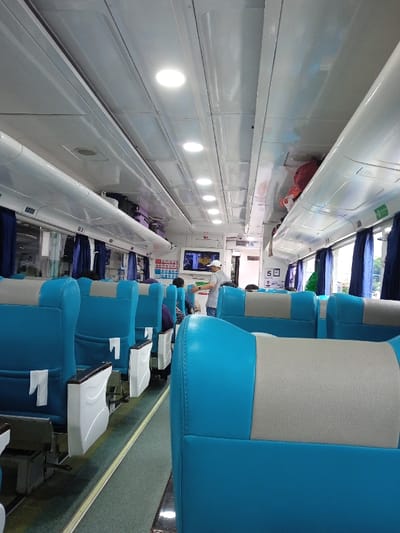 The image depicts the interior of a passenger train car, contextually situated within the city of Madiun, Indonesia. The cabin features a white, paneled ceiling uniformly lit by multiple recessed circular LED light fixtures. White, enclosed overhead luggage racks line both sides of the compartment, containing several items of luggage, including a visible red bag. Dark blue curtains cover the windows along both sides. The floor is carpeted in a greenish-grey tone, with a distinct yellow-green strip marking the central aisle.

Numerous high-backed passenger seats are arranged in pairs, separated by the aisle. Each seat is upholstered in a two-tone scheme, featuring light blue on the lower section and a light gray or beige on the upper section, complemented by white armrests. Seat-back pockets are visible, one containing a white rectangular insert.

Towards the forward section of the visible carriage, a male individual stands in the aisle, wearing a light-colored short-sleeved shirt and a white baseball cap. He is oriented towards the left side of the carriage (viewer's left). A flat-screen monitor is mounted on the wall to his right, displaying an image. Various informational signs, primarily white with blue text or graphics, are affixed to the cabin walls, including one prominently displaying the number "5." Multiple other passengers are partially visible, seated or obscured by the foreground seating. No specific dynamic interactions are discernible.