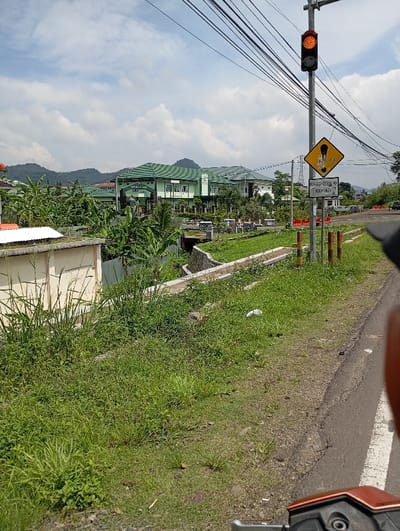 The scene is a roadside view in Kutawaringin, Indonesia, at coordinates -6.983 latitude and 107.533 longitude. A concrete drainage ditch with overgrown grass lies between the road and a row of green-roofed buildings. A traffic light with a red light is visible, mounted on a pole with power lines overhead. The road is paved and has a white line along the edge of the ditch. On the right side of the image, a yellow warning sign with a black exclamation mark is visible, with a second sign below in Indonesian. Vegetation and foliage are abundant, with a concrete wall and a few houses visible in the distance. The camera is positioned on the road looking towards the drainage ditch and buildings.