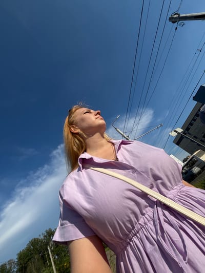 The image shows a young woman from the chest up, looking upwards against a clear blue sky. She has blonde hair and is wearing a light purple dress with short sleeves and a collared neckline. A white strap from a bag is visible across her chest. In the background, numerous power lines crisscross the sky, with a utility pole visible on the left. To the right, a tall building with multiple windows and a bus are partially visible, suggesting an urban environment. The low-angle shot emphasizes the vastness of the sky and the woman's upward gaze. The scene appears to be an outdoor public space in the city of Zavolzhye, Russia, likely during daylight hours.