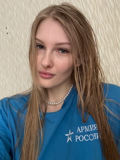 A young woman is captured in a headshot, seemingly lying down and looking upwards, though the image is rotated 90 degrees clockwise. She has long, straight, light brown hair that cascades around her head and shoulders. Her light-colored eyes, appearing greenish-blue, gaze directly at the viewer, and her lips are softly parted in a subtle, gentle smile.
She is wearing a light blue long-sleeved top. Across the chest area of her top, prominent white text is visible, which reads "АРМИЯ РОССИИ," translating to "ARMY OF RUSSIA." Below this text, a white five-pointed star emblem is also present. Around her neck, she wears a single strand of small, spherical pearl beads.
The background is a light beige or off-white surface with a subtle, wavy texture, resembling wallpaper or a patterned bedspread. The lighting is soft and even, suggesting an indoor setting without harsh shadows. The overall impression conveyed is one of calm and youthfulness. The location context provided is Кондратово, Russia.