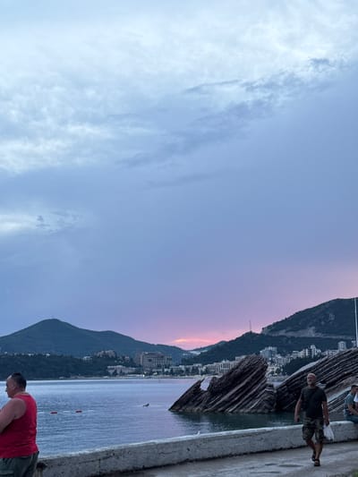 The image captures a serene evening scene in Rafailovići, Montenegro. The sky is a canvas of soft hues, transitioning from a pale blue on the left to a delicate blend of pink and purple on the right, indicating sunset or twilight. Wispy clouds drift across the sky, adding texture to the twilight atmosphere.
The foreground features a concrete walkway or embankment, with a rough texture and a slight sheen of water. To the right of the walkway, the calm sea reflects the colors of the sky. In the water, a few red and white buoys are visible, marking a swimming area.
On the left side of the image, a lush green mountain slopes down towards the sea. Nestled on the mountainside is a cluster of buildings, appearing to be hotels or apartments, their lights beginning to twinkle as the day fades. The architecture suggests a coastal resort town.
Several people are present. In the lower right, a man in a red tank top and dark shorts is captured in a bent-over posture, possibly resting or engaged in an activity near the water's edge. Further up the walkway, another man in a dark t-shirt and camouflage shorts stands, looking towards the sea and the distant landscape. A partial view of a third person can be seen in the upper right, near the edge of the frame.
Dominating the mid-ground are several large, abstract, wooden structures, with a rough, layered texture, resembling driftwood or natural formations. These structures add a unique artistic or natural element to the scene.
The overall mood is peaceful and contemplative, with the natural beauty of the landscape enhanced by the soft evening light and the presence of human activity. No visible text is present in the image.