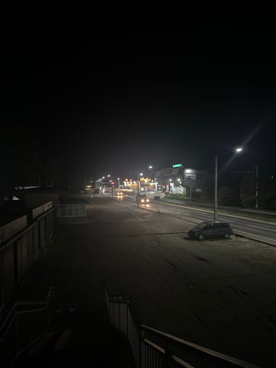 The image depicts a nighttime street scene in Strășeni, Moldova. The foreground shows a dark, paved area with a grey, four-door hatchback car parked on the right side. A metal fence and stairs are visible on the left. The midground features a road with visible lane markings, illuminated by streetlights and the lights from businesses on the right. Several cars are in motion on the road, their headlights and taillights creating streaks of light. A few buildings with illuminated signage are present along the roadside, including one that appears to be a technology store. The background consists of dark trees and foliage, suggesting a less developed area beyond the immediate urban environment. The overall atmosphere is one of a quiet, somewhat desolate, urban fringe at night.