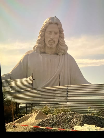 The image shows a large, white statue of Jesus Christ with outstretched arms, towering over a construction site. A small bird is perched on top of the statue's head. In the foreground, a corrugated metal fence blocks the view of the statue's base, and piles of sand and gravel are visible behind it, along with construction tape. The statue is located in the city of Dilijan, Armenia, and appears to be part of a larger religious or tourist attraction. The sky is overcast with wispy clouds, and there is a faint rainbow visible in the upper left corner of the image. The overall scene suggests ongoing construction or development around the statue.