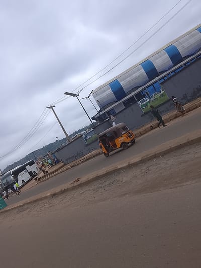 This media file captures a street scene in Enugu, Nigeria, under an overcast sky. The perspective is from a slightly elevated angle, looking down onto a moderately busy road.
The primary subjects are various vehicles and a few people. On the left, a white bus with "IMESE" and "CRUZ" visible on its side is parked. Several people, some wearing bright yellow vests, are seen near the bus, suggesting a possible bus station or transport hub. Further down the road, a yellow three-wheeled auto-rickshaw, commonly known as a 'keke', is in motion. Two individuals are walking on the sidewalk to the right of the road, with one person in a dark suit and the other in a patterned dress. Behind them, a large building with a distinctive blue and white striped roof is visible. A large sign with images of white vans is mounted on the building's dark grey wall.
The scene appears to be during daylight, though the cloudy weather obscures direct sunlight, creating a diffused light. The road is paved with asphalt, and there are raised concrete curbs and a dirt shoulder. The background features a gentle slope covered in greenery, with buildings scattered on the hillside, typical of an urban environment with natural topography. The overall atmosphere suggests a typical day with ongoing commercial and transportation activities. No specific event is clearly depicted, but the presence of the bus and the keke indicates movement and daily routines.
