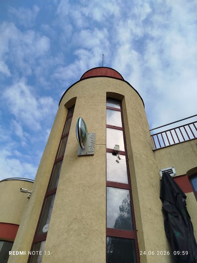 The image captures a low-angle view of a cylindrical building with a red dome-like roof, set against a cloudy blue sky. The building's exterior is textured and painted in a light yellow color, with dark red window frames. A prominent oval sign with a green and white logo, appearing to be for "Heineken," is mounted on the building. Several tall, narrow windows are visible. A security camera is mounted on the left side of the building, and a black tarp or umbrella is draped over a vent on the right side. A metal railing with red posts runs along a higher section of the building. The timestamp "24.06.2026 09:59" and the text "REDMI NOTE 13" are overlaid on the image. The context provided indicates this location is in Vratsa, Bulgaria.