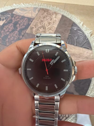 A person's hand holds a silver-colored metallic wristwatch with a black face, prominently displaying it against a patterned fabric background. The hand, with a light-to-medium skin tone, wraps around the watch, showing the thumb, index, and middle fingers.
The watch features a black dial with silver-colored rectangular hour markers and small white minute markings around the edge, with numbers visible at intervals of five minutes (e.g., '05', '10', '15'). The brand name "HUGO" is displayed vertically in red text on the left side of the watch face. On the right side of the face, stacked text reads "3 BAR" and "30 METERS" in white, indicating water resistance. The hour and minute hands are silver-colored, while the thin second hand is red. The watch case and bracelet are made of polished silver-colored metal, with a knurled crown visible on the right side of the case.
The background consists of an ornate rug or textile with a variegated pattern, including sections of beige, light brown, and darker gray-blue with intricate floral and abstract motifs. A light-colored fringed edge of the textile is visible on the left side of the frame. The lighting suggests an indoor setting. The image captures the watch in detail, indicating its display or presentation in Dragor, Bulgaria.