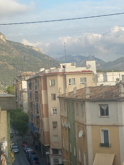The image depicts a view of buildings and mountains in Gandia, Spain. In the foreground, a portion of a building with a gray, textured facade is visible on the left. Adjacent to it, several apartment buildings with various architectural styles and colors are lined up, extending into the mid-ground. These buildings have multiple floors, windows, and balconies. Some of the buildings feature awnings, satellite dishes, and air conditioning units. A street runs alongside these buildings, with several cars parked along its edge and a few in motion. Trees are visible on the left side of the street. In the background, a range of mountains with green, forested slopes rises against a sky with scattered clouds. Power lines stretch across the upper part of the image. The lighting suggests it is daytime, possibly late afternoon, given the shadows.