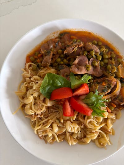An overhead shot showcases a white plate filled with a hearty meal. The dish consists of a savory stew of sliced meat and peas in a rich, red sauce, served alongside a generous portion of pasta. The pasta is a mix of short, curved macaroni and thin vermicelli noodles, all coated in a light sauce. Garnishing the dish are fresh green lettuce leaves and wedges of bright red tomato, adding a pop of color and freshness. The entire plate is set against a clean, light-colored countertop, hinting at a domestic setting, possibly a kitchen or dining area. The lighting suggests it might be daytime, with natural light illuminating the scene. The image does not depict any specific landmarks or indicate a location in Miami, United States of America.