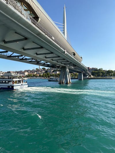 This image captures a view of the Golden Horn Metro Bridge in Istanbul, Turkey, from the water. A large, modern bridge with a light grey, curved structure dominates the upper portion of the frame, supported by thick concrete pillars. The bridge appears to be elevated, with visible metal supports beneath it. On the left side of the image, a white ferry with blue trim is moving through the water, creating a noticeable wake. The ferry has "METE II ISTANBUL" written on its side. In the background, across the water, is a dense cityscape of buildings with red-tiled roofs, interspersed with trees and a tall minaret, suggesting a historic district. The water is a vibrant turquoise color, with visible ripples and some white foam from the ferry's passage. The sky above is a clear, bright blue with a few small birds flying in the distance. The overall impression is a sunny day with active maritime and urban life.