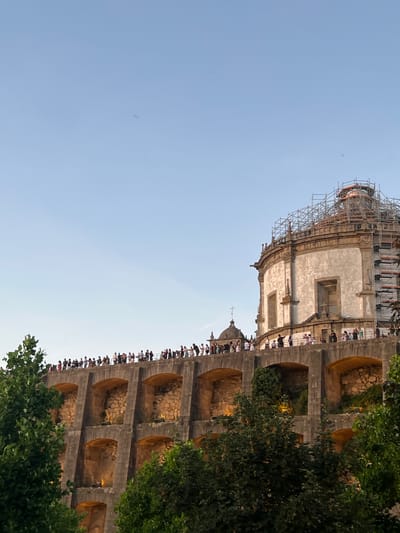 The image shows the Clérigos Church and its bell tower, a prominent landmark in Porto, Portugal, seen from across the Douro River in Vila Nova de Gaia. The church is partially covered in scaffolding, indicating ongoing renovation work. A crowd of people are visible on the top of a wall in the foreground, looking out at the view. Lush green trees frame the bottom of the image. The sky is a clear, pale blue, with a few faint streaks that could be clouds or birds. The architectural style of the church is Baroque, characterized by its dome and elaborate facade. The wall in the foreground appears to be part of a garden or park, with arched openings revealing illuminated interiors. The scene suggests a popular tourist spot, with many visitors gathered to admire the view and the historic architecture.