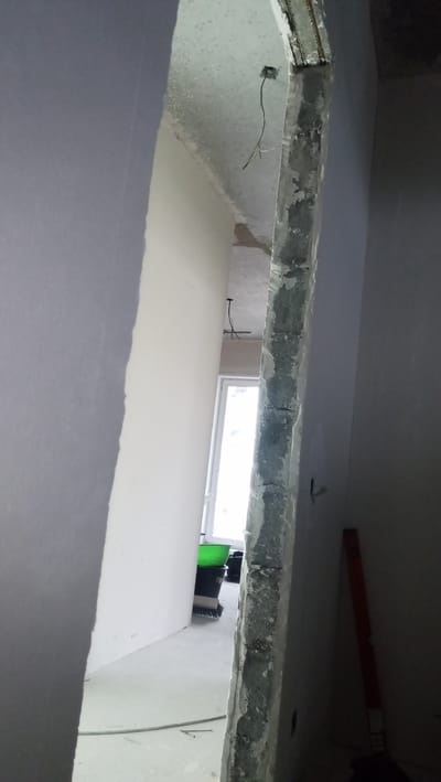 The image depicts an interior construction site, likely a residential apartment undergoing renovation, in Vladivostok, Russia. The viewpoint is from a doorway or opening, looking into a hallway or room.
On the left, a newly plastered wall is visible, with a rough, unfinished edge where it meets the opening. The wall is painted or primed in a light gray color.
Dominating the center of the image is a rough, unfinished concrete pillar or wall section, showing exposed rebar at the top. This structure appears to be a structural element of the building and has a textured, grey, and somewhat damaged surface.
To the right of the concrete pillar, another wall is visible, painted in a dark grey or black color, suggesting it might be a finished surface or a different material. An orange spirit level is leaning against this wall.
In the background, a window with a white frame is letting in natural light. In front of the window, there are several construction-related items: a bright green plastic bucket, a black bucket, and what appears to be a broom. The floor is concrete and appears dusty and unfinished. Electrical wires are hanging from the ceiling in the opening. The overall impression is of an active construction or renovation zone.