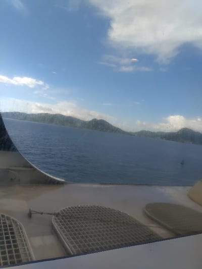 The scene is a view from a ferry, looking out at the sea. The coordinates, 10.63 latitude and -61.566 longitude, place the location in the Port of Spain, Trinidad and Tobago. In the distance, a mountain range appears on the horizon. The sky is blue with a few white clouds. The ferry deck is visible in the foreground, with two round, square-grid ventilation panels. The sea is a light blue and appears calm. A lone boat is in the distance. The photo was taken during the daytime.