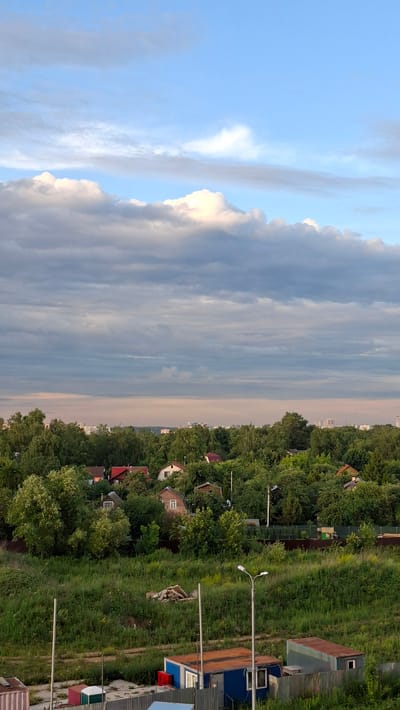 This is a vertical, eye-level, outdoor shot of a rural or suburban landscape in Vidnoye, Russia. The image is split roughly in half, with the left side dominated by a bright blue sky filled with wispy, grey, and white clouds, suggesting it is either early morning or late afternoon. The right side of the image depicts a dense line of green trees bordering a grassy field with several small houses and outbuildings scattered throughout. The buildings have red and blue roofs, and some appear to be constructed from wood or corrugated metal. Two lampposts are visible, one in the foreground and another further back. The overall mood is serene and tranquil, with the natural elements of the sky and foliage creating a sense of peace. There is no visible text in the image.