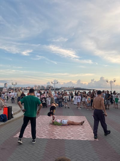 The image captures a street performance on a waterfront promenade in Gelendzhik, Russia. The sky is a pale blue with wispy white clouds, suggesting it is either early morning or late afternoon. In the foreground, two individuals are engaged in a breakdancing routine. One person is performing a handstand, balanced on their hands with their legs extended upwards. The other person is lying on their back on a patterned mat, with their head facing towards the viewer and their legs extended. A third individual, wearing a green t-shirt and black pants, stands facing away from the viewer, observing the performance. To the right of the performers, another shirtless man in dark pants walks away from the scene.

A crowd of onlookers is gathered around the performers, creating a lively atmosphere. The promenade is paved with interlocking bricks in a red and grey pattern. In the background, the Gelendzhik cityscape is visible, with a cluster of buildings and architecture along the coast. Beyond the city, the sea stretches out to the horizon, dotted with boats. Streetlights and lampposts line the promenade, adding to the urban feel of the scene. The overall impression is one of a public gathering, with entertainment and leisure activities taking place by the sea.